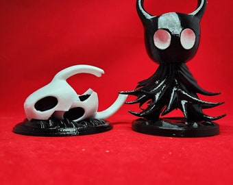 May include: Two detailed figurines, one black and one white, sit on a red surface. The black figure has a horned head and a flowing cape. The white figure resembles a skull with a curved horn. Both figures are on black bases.