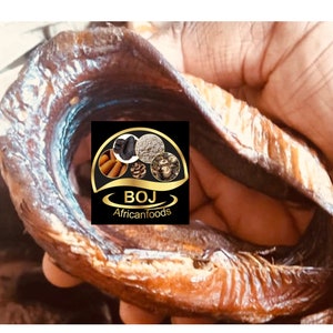 May include: Close-up of a dried fish, likely a catfish, with a golden brown color. The fish is held in a hand, and the background is blurred. A logo for "BOJ Africanfoods" is visible in the lower right corner.