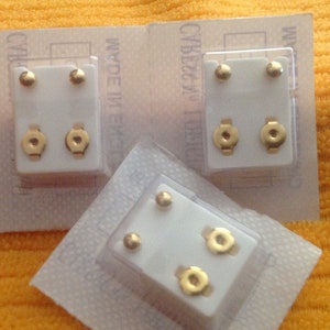 May include: Three clear plastic packages containing pairs of gold-colored stud earrings. Each package has the text "MADE IN ENGLAND" printed on it.