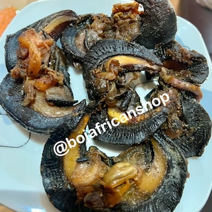 May include: Close-up of a plate of cooked snails. The snails are black and have a brown, meaty interior. The snails are arranged in a circular pattern on the plate. The text '@bojafricanshop' is visible in the lower left corner of the image.
