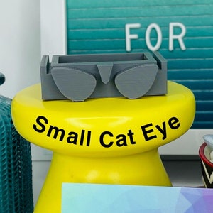 Op de afbeelding: Een kleine grijze plastic sjabloon voor het maken van kattenoogbrillen voor poppen. De sjabloon staat op een gele standaard met de tekst "Small Cat Eye" erop gedrukt. Er is een visitekaartje met de tekst "Stitches & Whimsy Handmade" erop. Er zijn ook twee tangen en een kleine potplant met een succulent.