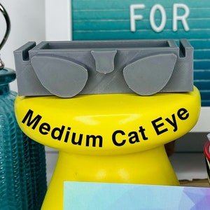 Op de afbeelding: Een gele plastic mal voor het maken van poppenogen, met het label "Medium Cat Eye". De mal ligt op een houten oppervlak met een visitekaartje waarop "Stitches & Whimsy Handmade" staat.