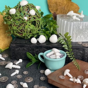 Puede incluir: Una colección de ojos de seguridad de plástico blanco para manualidades, algunos están en una taza pequeña de color turquesa, otros están en una superficie de madera y otros están en una superficie negra con un libro y una roca blanca.