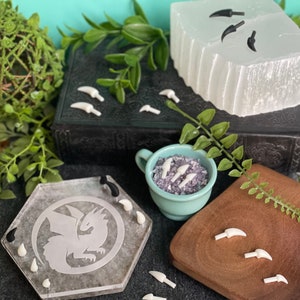 May include: A set of white and black dragon claw charms. The charms are displayed on a variety of surfaces, including a wooden tray, a clear glass tray with a dragon design, and a white selenite crystal. The charms are perfect for adding a touch of fantasy to your jewelry or crafts.