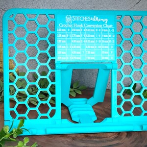 May include: A turquoise blue crochet hook holder with a honeycomb pattern. The holder has a chart with crochet hook sizes and corresponding measurements in millimeters and inches. The chart is labeled "Crochet Hook Conversion Chart" and "STITCHES Whimsy".