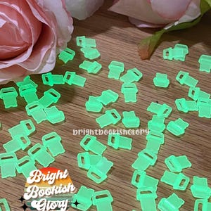 May include: A large quantity of bright green glow-in-the-dark plastic slider buckles. The buckles are scattered on a wooden surface. The text "Bright Bookish Glory" is visible in the lower left corner of the image.