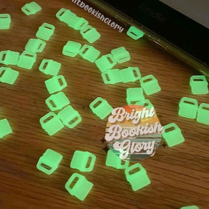 May include: Glow-in-the-dark Kindle page holders scattered on a wooden surface. The neon green holders are rectangular with a small handle. A Kindle device is partially visible. The text "brightbookishGLORY" and "Bright Bookish Glory" are also visible.