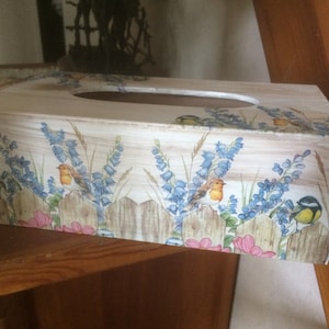 May include: A rectangular wooden tissue box with a design of birds, blue flowers, and a wooden fence. The box has a distressed finish, and is placed on a wooden shelf. The measurements are in centimetres.