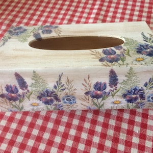 May include: A wooden tissue box cover with a floral design. The design features purple irises, white daisies, and green foliage on a white background.