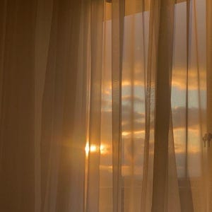 Può includere: Tende trasparenti e chiare sono appese davanti a una finestra, con il sole che splende attraverso. La luce del sole crea un bagliore dorato e caldo, illuminando le tende e il cielo visibile attraverso la finestra. La scena evoca pace e tranquillità.