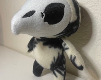 May include: A small, white and black plush toy with a skull-like head and a long beak. The toy has a black and white patterned body and is standing on a white surface.