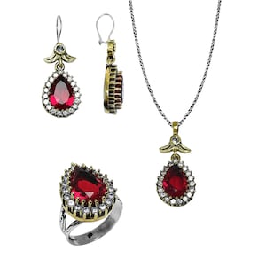 Op de afbeelding: Een sieradenset met een ring, oorbellen en een ketting. Elk stuk heeft een druppelvormige, dieprode edelsteen omringd door kleine, heldere stenen. Het metaal heeft een goudkleurige afwerking en de ketting heeft een zilveren ketting.