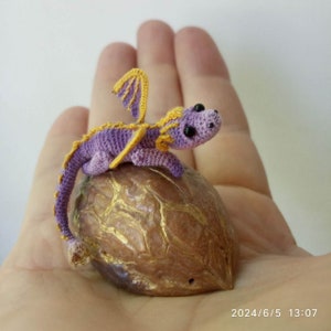 May include: A miniature crocheted purple dragon with yellow wings and a yellow spine crest, perched atop a brown walnut shell that features gold-painted cracks. The dragon has small black bead eyes and a long, slender, curled tail.