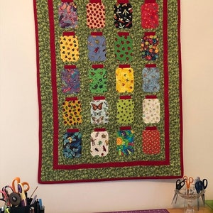 以下が含まれることがあります： さまざまな昆虫や花の模様が施された、瓶の形をした16個のパッチが特徴の手作りのキルト。キルトは緑色の背景に赤い縁取りがあり、白い壁に掛けられています。キルトの幅は約100cm、長さは127cmです。
