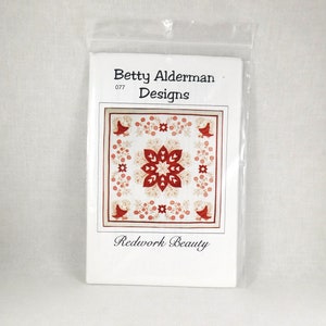 May include: A white paper pattern packet with red text that reads "Betty Alderman Designs" and "Redwork Beauty". The pattern features a red and white quilt block with birds and flowers.