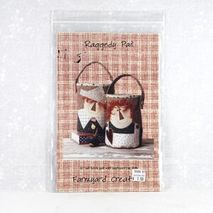 May include: A sewing pattern packet titled Raggedy Pail and Farmyard Creats featuring two fabric pails with appliqued rag doll faces. The pattern cover displays a red and white grid background, text, and a photo of the finished fabric pails.