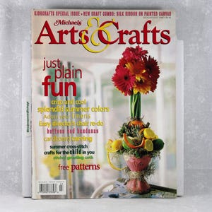 May include: A July/August 1997 Michaels Arts & Crafts magazine issue featuring a floral arrangement with red and orange gerbera daisies in a pink vase decorated with citrus slices and greenery. The cover includes text such as just plain fun.
