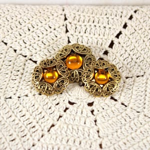 May include: Three circular buttons featuring an intricate, antiqued gold-tone metal filigree frame surrounding a smooth, translucent amber-colored cabochon center. The buttons are arranged in a cluster, showcasing detailed metalwork patterns.