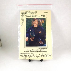 May include: A pattern for a fleece jacket applique with a "Camp Peek-A-Boo" theme. The pattern features a blue jacket with a moon, trees, and a cabin applique. The pattern is from Creative Uniques, Inc. and includes handmade stoneware buttons.