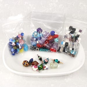 May include: An assortment of colorful beads in clear plastic bags and on a white ceramic dish. The beads include various shapes, sizes, and colors, such as blue, pink, red, and black. Some beads have unique patterns and textures, perfect for jewelry making.