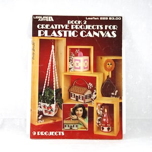 May include: A vintage book cover titled "Creative Projects for Plastic Canvas" by Leisure Arts. The cover features a red background with wooden display boxes showcasing various craft projects, including a hanging planter, a miniature house, and a candle holder.