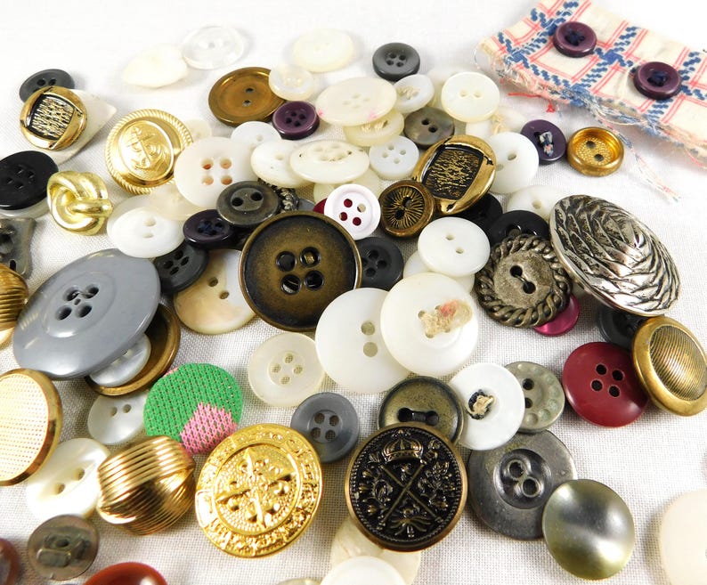 May include: A collection of vintage buttons in various colors, shapes, and sizes. The buttons are made of different materials, including metal, plastic, and fabric. Some of the buttons have intricate designs, while others are simple and plain.