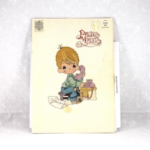 May include: A vintage "Precious Moments" cross-stitch pattern book. The cover features a cross-stitched illustration of a young boy on the phone. The title "Precious Moments" is in a decorative script. The book is cream-colored with some wear.