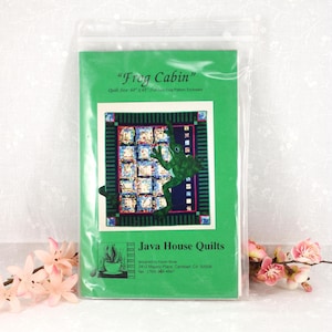 May include: A green package with a quilt pattern titled "Frog Cabin." The quilt design features a frog, squares, and a striped border. The package also includes the text "Java House Quilts."