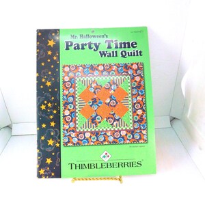 May include: A book titled "Mr. Halloween's Party Time Wall Quilt" with a green cover and a colorful quilt design. The quilt features orange, blue, and green patterns. The book is propped up on a gold-colored stand.