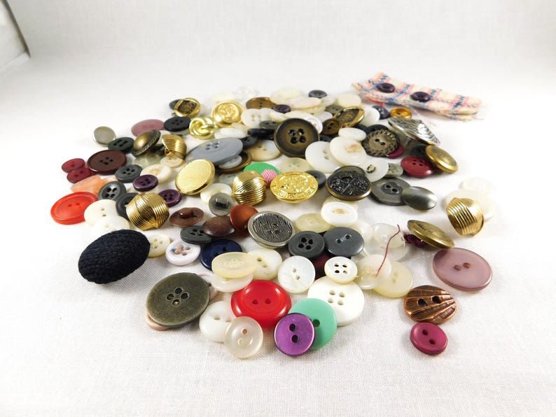 May include: A large collection of assorted buttons in various colors, shapes, and sizes. The buttons are made of plastic, metal, and fabric. Some buttons have a shiny finish, while others have a matte finish.