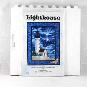 May include: A craft kit featuring a lighthouse design. The image shows a white lighthouse with a red roof, set against a blue sky with a sunburst effect. The text "Lighthouse" is at the top.