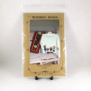 May include: A craft pattern titled "Bloomin' Minds" in a clear plastic sleeve. The pattern features a light green sweater with daisy designs, a daisy-themed quilt, and the text "Pushin' Up Daisies #168". The pattern is displayed on a wooden bench.