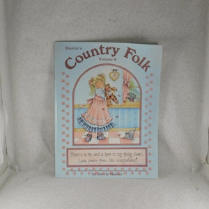 May include: A book titled "Darcie's Country Folk Volume 6" with a colorful illustration of a girl holding a teddy bear. The book includes step-by-step embossing instructions and a color worksheet.