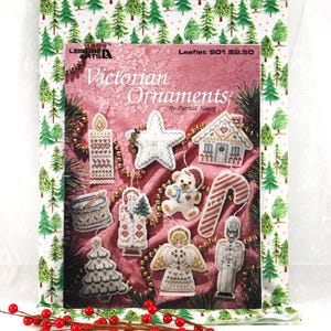 May include: A book titled "Victorian Ornaments" featuring cross-stitch designs of Christmas ornaments. The cover is framed by a fabric with green Christmas trees. A sprig of red berries is in the foreground.