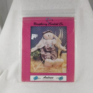 May include: A sewing pattern for a 24 inch plush bunny rabbit named Andrew. The pattern is from Raspberry Cordial Co. and features a bunny wearing a blue and white shirt, brown pants, and a white hat.