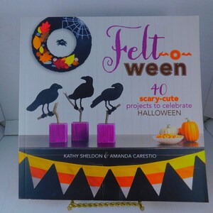 May include: Book cover with the title "Felt Ween" and the subtitle "40 scary-cute projects to celebrate HALLOWEEN". The cover features a felt wreath with a spider, three black felt crows, and a felt candy corn garland.