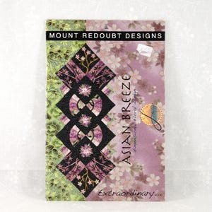May include: A book cover for "Asian Breeze" by Mount Redoubt Designs. The cover features a geometric design with black, purple, and green patterns. The background is a floral print in shades of pink and white. The word "Extraordinary..." is at the bottom.