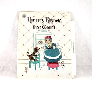 May include: A cross-stitch design of a book titled "Nursery Rhymes that Count." The cover features a scene with a woman, a dog, and a mouse. The woman is wearing a teal dress and a pink hat. The dog is sitting on a teal chair.