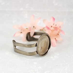 May include: A silver-toned metal bracelet with an oval bezel setting. The bracelet has a split band design and is displayed with pink flowers in the background. The bezel is designed to hold a cabochon or other decorative element.