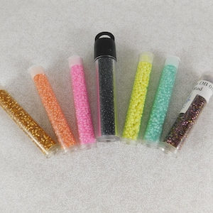 May include: A collection of small, colorful beads in clear tubes. The beads are in various colors, including gold, orange, pink, black, yellow, teal, and a reddish-brown. The tubes are arranged in a semi-circle.