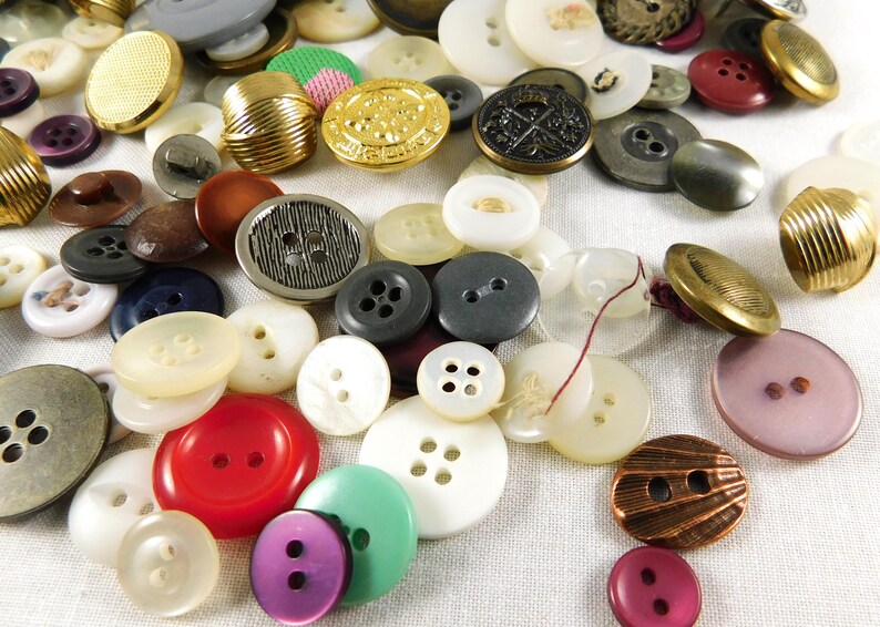 May include: A collection of assorted buttons in various colors, shapes, and sizes. The buttons are made of different materials, including metal, plastic, and fabric. Some buttons have a decorative design, while others are plain.