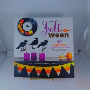 May include: Book cover with the title "Felt-oween" and the subtitle "40 scary-cute projects to celebrate HALLOWEEN". The cover features a felt wreath with a spider, three black crows, and a pumpkin.