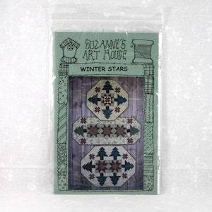 May include: A package featuring a quilt pattern titled "Winter Stars" by Suzanne's Art House. The pattern includes three designs with geometric shapes, stars, and trees in shades of blue, red, and green on a wooden background. The package is in a clear plastic bag.