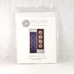 May include: A package featuring a stained glass applique pattern. The design includes two vertical panels with star and geometric patterns in shades of purple, blue, and brown. Text on the package reads "STARFIRE BANNER" and "THREE SWANS STUDIOS."