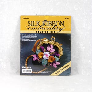 May include: A Bucilla Silk Ribbon Embroidery Starter Kit for Carnations, model 41831. The packaging features a black purse with gold hardware and colorful ribbon flowers. Text includes Silk Ribbon Embroidery Starter Kit, Carnations, and 41831.
