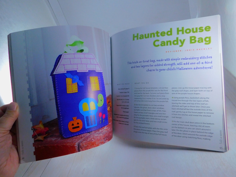 May include: A craft project for a haunted house candy bag. The bag is made of felt and features a purple house with a skull, pumpkin, and bat appliques. The instructions are written in a book.