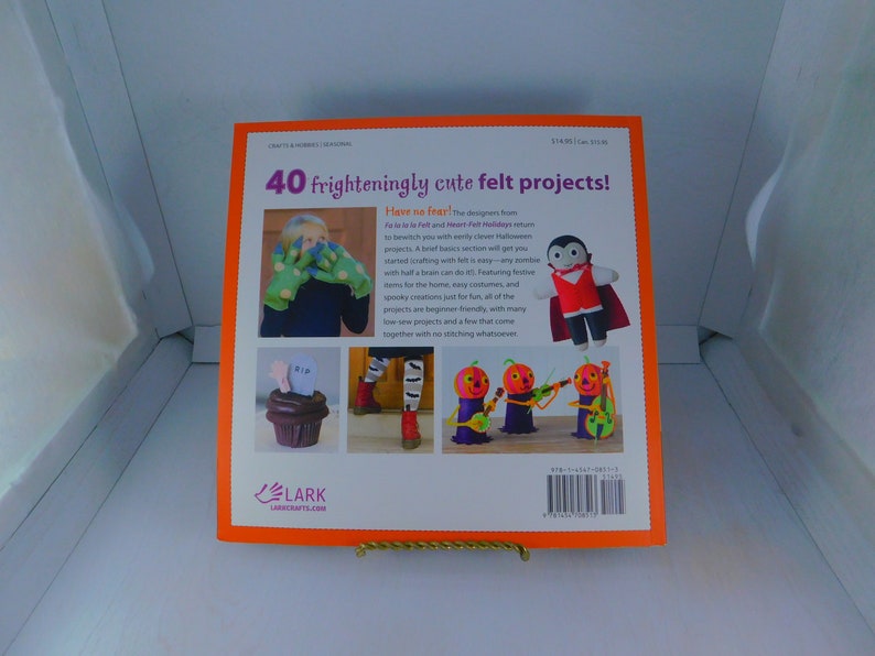 May include: The back cover of a book titled "40 frighteningly cute felt projects!"  The book features a bright orange background with a variety of felt projects, including a vampire, a ghost, and a cupcake. The book is published by Lark Crafts.