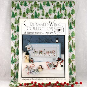 May include: A framed cross-stitch pattern book titled "Crossed Wings Collection: A Frosty Feast No. 29" featuring snowmen. The book is surrounded by fabric with a green tree pattern and a sprig of red berries.