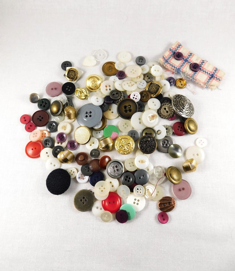 May include: A large collection of assorted buttons in various colors, sizes, and materials, including plastic, metal, and fabric. The buttons are arranged in a random pattern on a white surface.