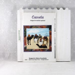 May include: A package for machine appliqué featuring a desert scene with camels. The cover reads "Camels" and "designs for machine appliqué." Designed by Debora Konchinsky. The package is in a clear plastic sleeve.